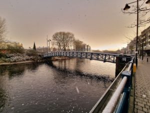 The Bridge of Light, a footbridge over Sligo's serene River Garavogue, in the snow. The light is a soft sepia gold, with the clouds verging on purple and sunrise making a white streak in the sky in the background.