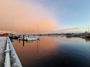 The Sligo docks in a light snow, with sunrise coloring a cloud over half the sky rose gold, and the rest of the sky pale morning blue. The colors are reflected in the almost-perfectly-still water.